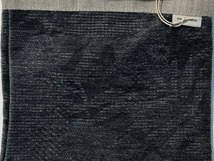 En mørk struktureret stofprøve med en lysegrå topkant, en metalring i hjørnet og en lille hvid etiket.