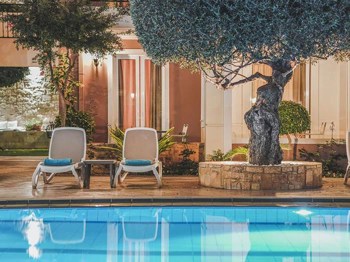 To liggestole med blå håndklæder står ved siden af en klar swimmingpool i skyggen af et stort træ foran en bygning med glasdøre og varm belysning.