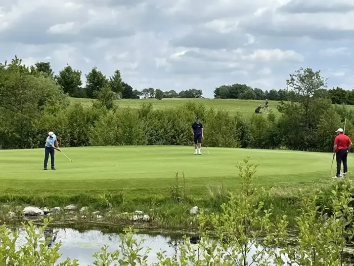 Tre personer spiller golf på en grøn bane omgivet af træer og buske under en overskyet himmel.