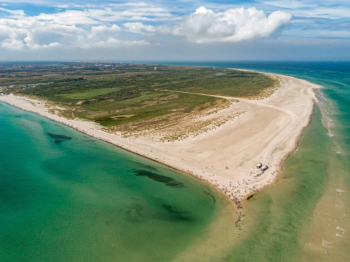 Luftfoto af en sandhalvø, der strækker sig ud i turkisgrønt havvand, med mennesker på stranden og græsarealer i baggrunden under en delvist overskyet himmel.