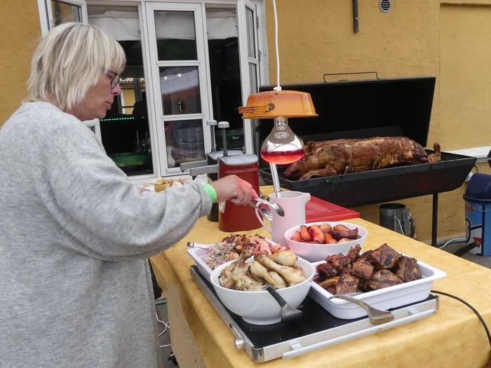 En person serverer mad fra et buffetbord med stegt kød, kartofler, grøntsager og en helstegt gris i baggrunden.