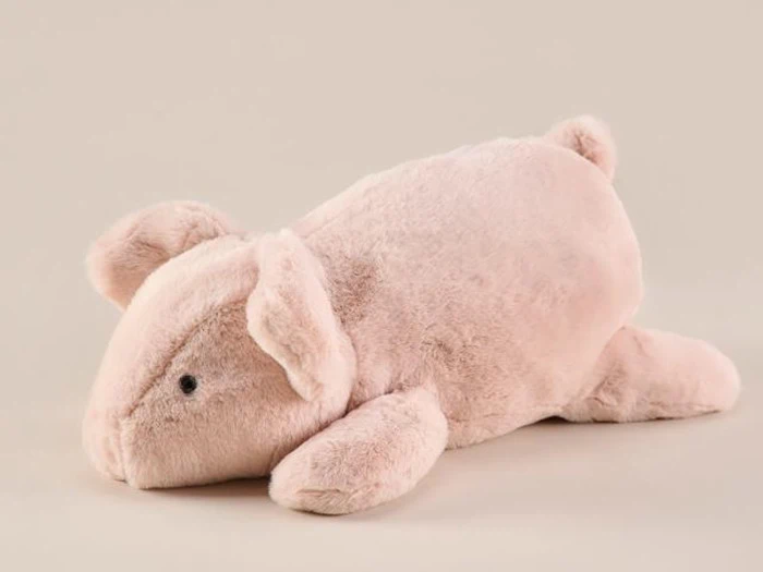 En blød, lyserød plysgris ligger på maven mod en almindelig beige baggrund.