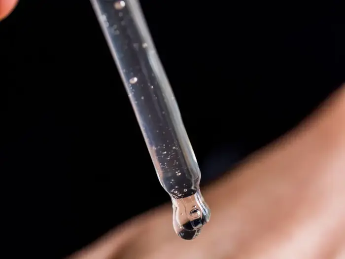 Et nærbillede af en glasdråbe, der frigiver en klar væske med små luftbobler indeni, mod en sløret, mørk baggrund.