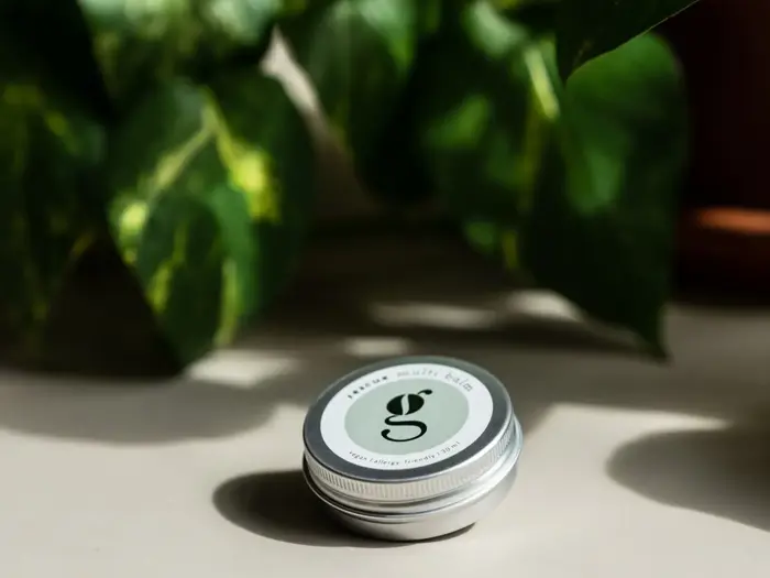 En lille rund dåse mærket "multi balm" med et lille bogstav "g" på låget, placeret på en lys flade med grønne bladplanter i baggrunden.