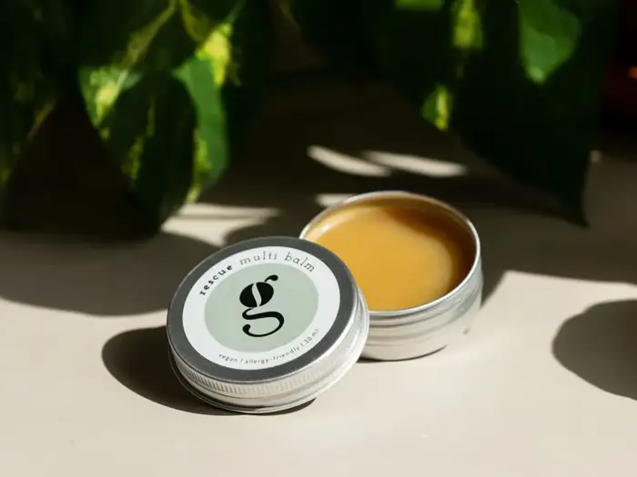 En dåse rescue multi balm med låget af ligger på en overflade og afslører balmen indeni. Grønne blade og sollys skaber skygger i baggrunden.