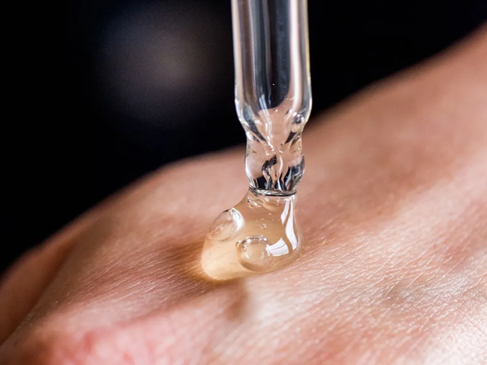 Et nærbillede af en dråbetæller, der doserer et klart serum på en persons håndryg, mod en mørk baggrund.