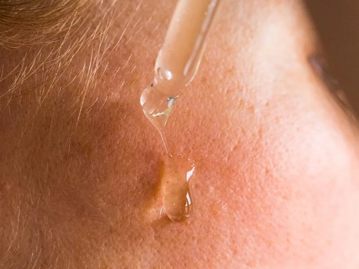 Et nærbillede af en dråbetæller, der påfører serum eller olie på en persons kind, viser hudens struktur og en klar dråbe.