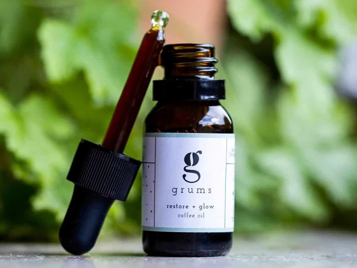 En lille brun glasflaske mærket grums restore + glow kaffeolie med en dråbetæller hvilende på flasken, sat op mod en sløret grøn bladbaggrund.