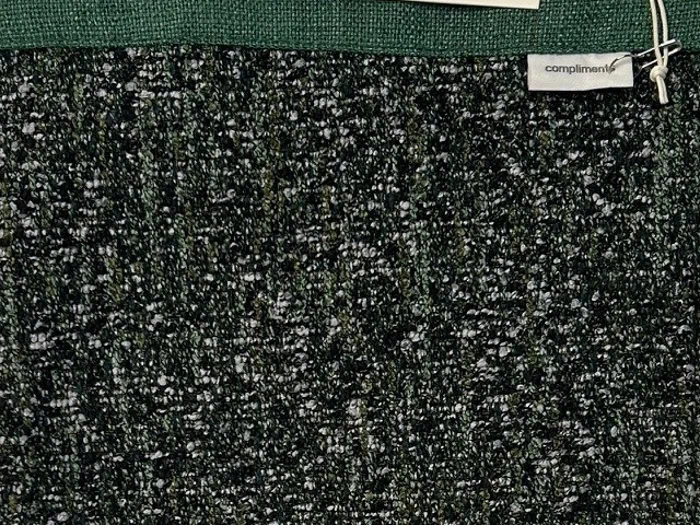 Nærbillede af et struktureret mørkegrønt og sort stof med et lille mærke, hvor der står kompliment i øverste højre hjørne.