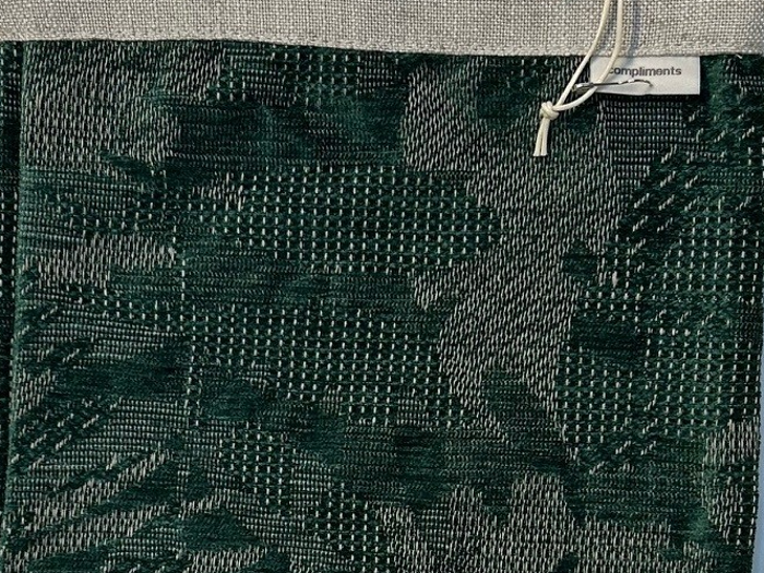 Et stykke mørkegrønt struktureret stof med abstrakte mønstre, fastgjort til en beige header med et mærke, hvor der står complimints, og en hvid snoresløjfe.