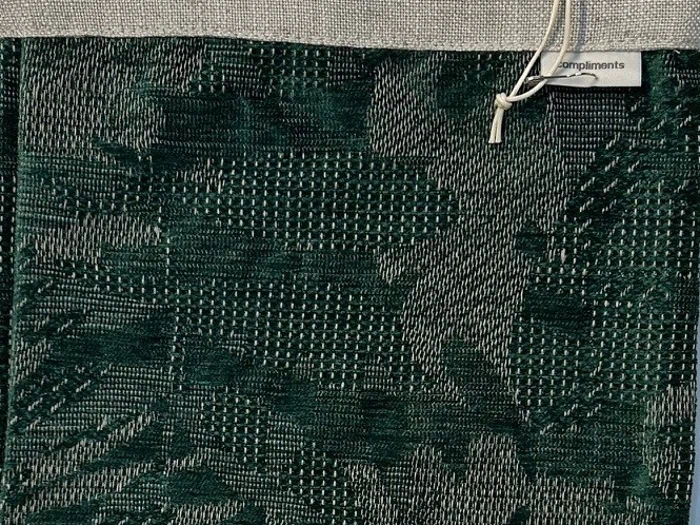 Et stykke mørkegrønt struktureret stof med abstrakte mønstre, fastgjort til en beige header med et mærke, hvor der står complimints, og en hvid snoresløjfe.
