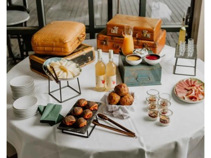 Et rundt bord med vintagekufferter, juiceflasker, kager, pålæg, fade, skåle med saucer og glas, dækket til en brunch- eller morgenmadsbuffet.