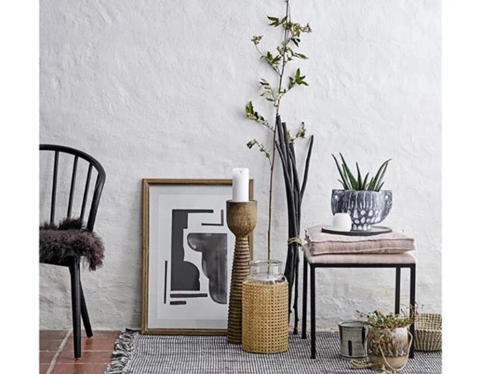 Minimalistisk interiør med en sort stol, et abstrakt indrammet kunstværk, en høj plante, en lysestage, en kurv, en potteplante, et sammenfoldet tæppe på en bænk og dekorative genstande på et tæppe mod