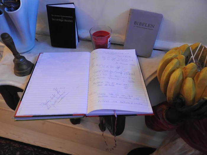 En åben gæstebog på et bord med håndskrevne indlæg, et lille rødt lys, to bøger og en klase bananer ved siden af.