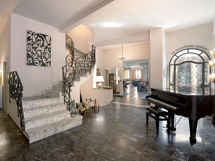 Elegant foyer med marmortrappe, udsmykket sort gelænder, sort flygel, stort vindue og moderne kunst på væggen. Rummet har polerede stengulve og varm belysning.