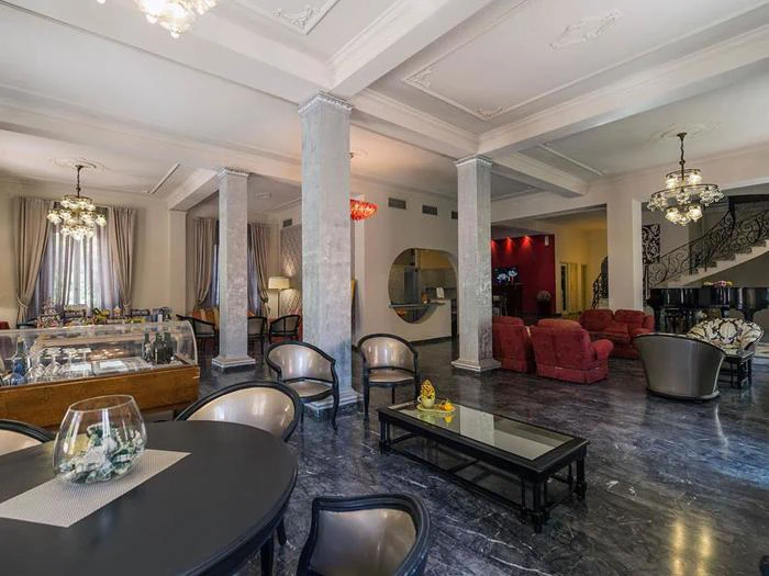 Elegant hotellounge med marmorgulv, runde borde, polstrede stole, glasindretning, lysekroner, røde lænestole og en trappe med udsmykkede gelændere i baggrunden.