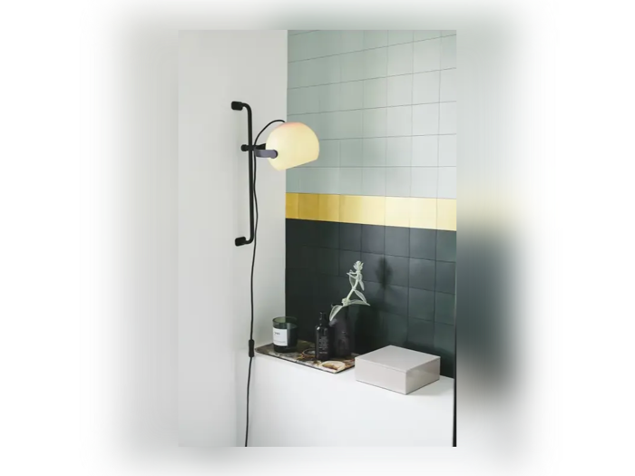 En moderne væglampe med en buet sort arm og en rund skærm er monteret over en afsats med dekoration mod grønne fliser med en gul stribe.