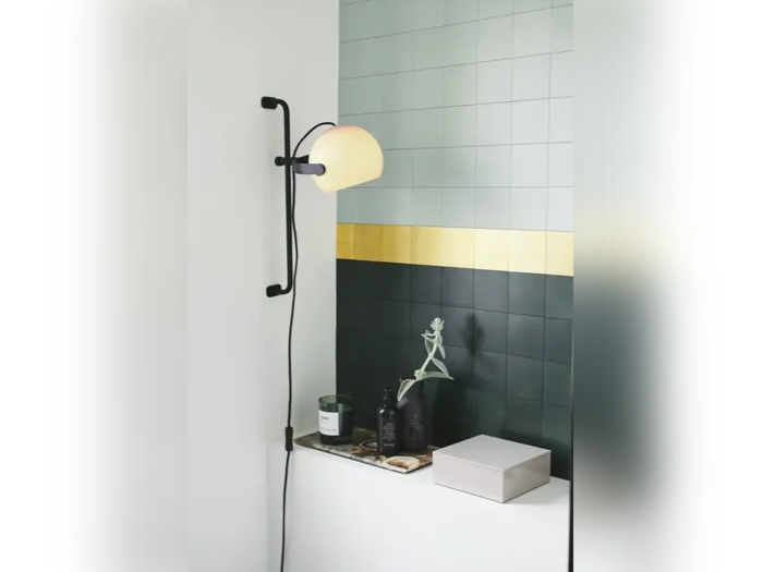 En moderne væglampe belyser en hylde med en plante, stearinlys og pynt; bagved ses en flisebelagt væg med grønne, gule og grå fliser.