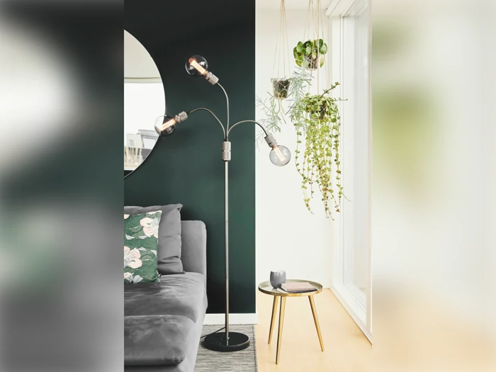 En moderne stue med en grå sofa, en sort gulvlampe, hængende planter, et rundt spejl og et lille rundt sidebord ved et lyst vindue.