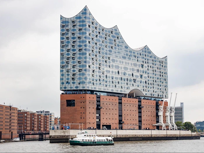En moderne glasbygning med bølget tag ligger på en murstensbase ved havnefronten, og en båd passerer foran.