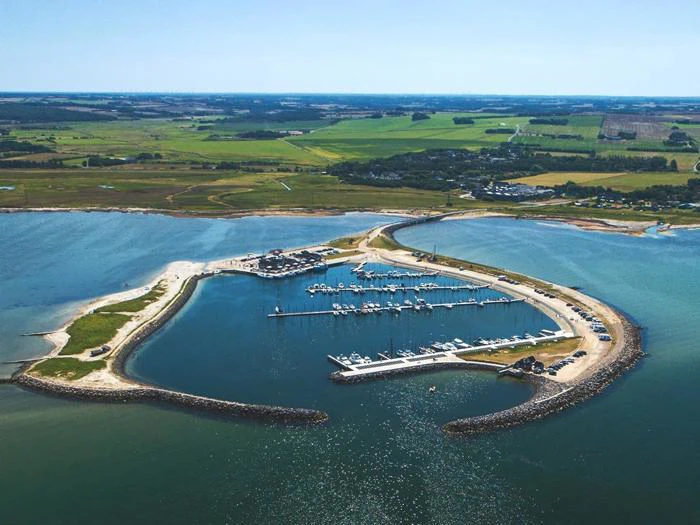 Luftfoto af en trekantet marina med dokker og både, omgivet af land, grønne marker og vand, under en klar blå himmel.