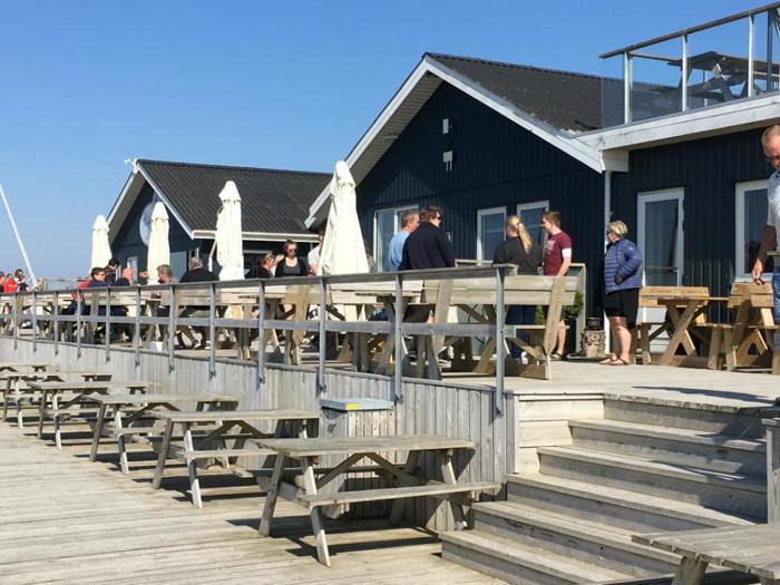 Folk står og sidder uden for en mørk træbygning ved havet med udendørs siddepladser og parasoller på en solrig dag.