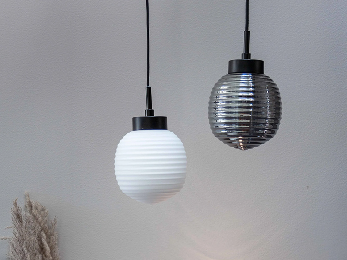 To pendler hænger side om side; den ene har en hvid, riflet glasskærm, og den anden har en mørkegrå, riflet glasskærm, begge med sorte beslag mod en lysegrå væg.