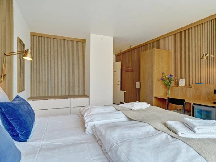 Moderne hotelværelse med en pænt redt seng, blå og hvide puder, foldede håndklæder, trædetaljer, et skrivebord med en stol og en vase med blomster.