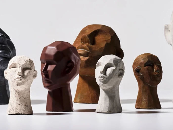 Flere stiliserede, facetterede skulpturer af menneskehoveder i forskellige størrelser og jordfarver vises på en almindelig, lys baggrund.