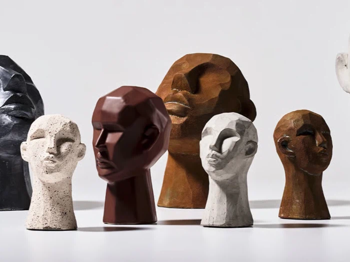 Syv skulpturelle, abstrakte menneskehoveder i forskellige størrelser og jordfarver er arrangeret sammen på en hvid flade med ensfarvet baggrund.