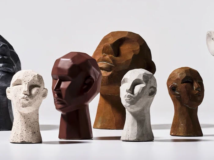 En gruppe skulpturelle, abstrakte menneskehoveder i forskellige jordfarver og teksturer er arrangeret på en glat overflade med en lys baggrund.