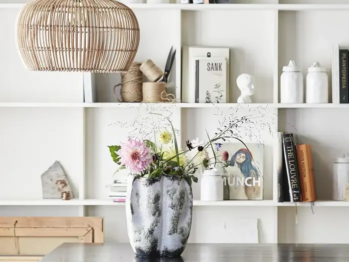 En keramikvase med lyserøde og gule blomster står på et bord foran en hvid bogreol fyldt med bøger, krukker og pyntegenstande. En vævet pendellampe hænger ovenover.