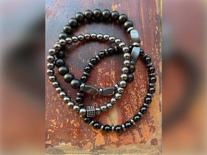 Tre perlearmbånd - et mat sort, et skinnende metallisk og et sort med en firkantet perle - er arrangeret på en rustik træoverflade.