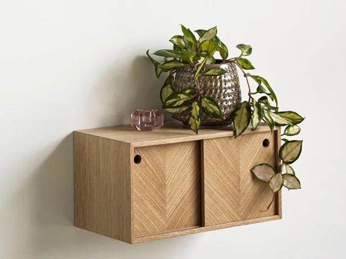 En lille væghylde af træ med skydelåger, der rummer en glasvase med en bladplante og en lille lyserød pyntegenstand på toppen.