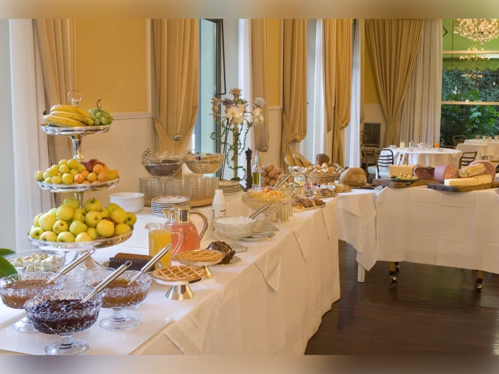 Et buffetbord med diverse frugter, kager, brød, morgenmadsprodukter, syltetøj og juice i en elegant spisestue med store vinduer og gule gardiner.