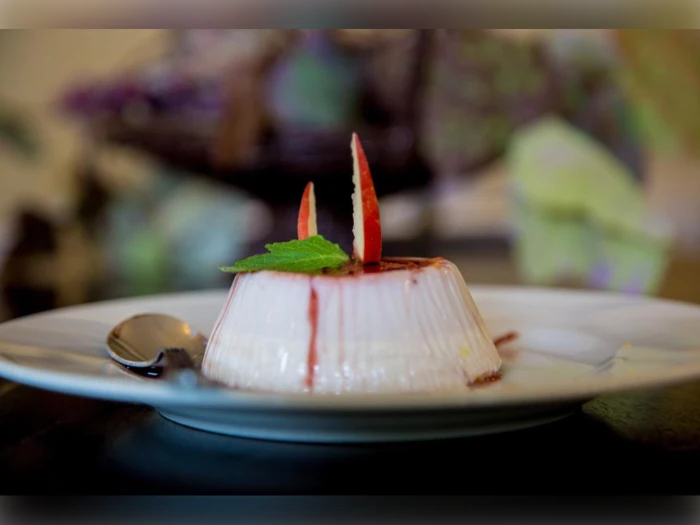 En cremet dessert toppet med rød sirup, æbleskiver og et mynteblad ligger på en hvid tallerken med en ske ved siden af.