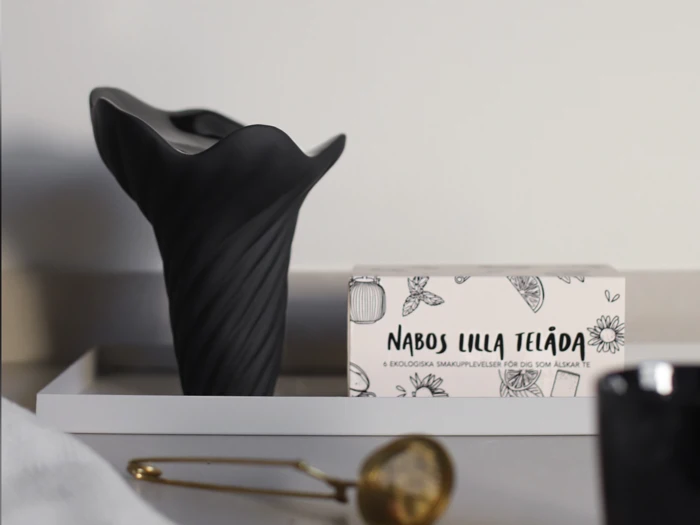 En sort vase og en æske med påskriften »Nabos Lilla Telåda« står på et hvidt fad ved siden af en gylden tefilter på en lys overflade.