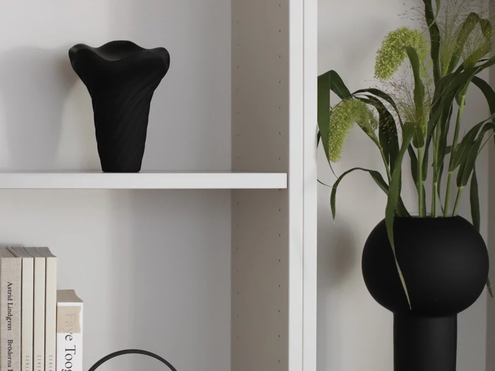 En sort vase på en hvid hylde, bøger stablet nedenunder og en sort, rund vase med grønt løv til højre.