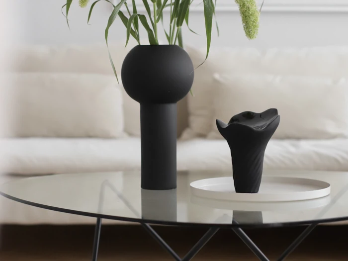 På et moderne sofabord af glas står en høj, sort vase med grønne planter og et mindre, skulpturelt, sort objekt på et hvidt fad, med en beige sofa i baggrunden.