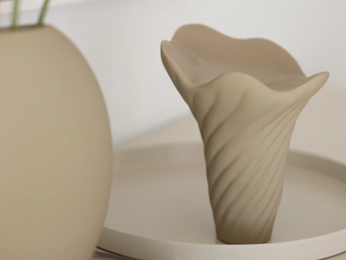 En beige, abstrakt keramisk vase med et bølget, snoet mønster står på et rundt fad ved siden af et glat, rundt objekt i en minimalistisk opstilling.