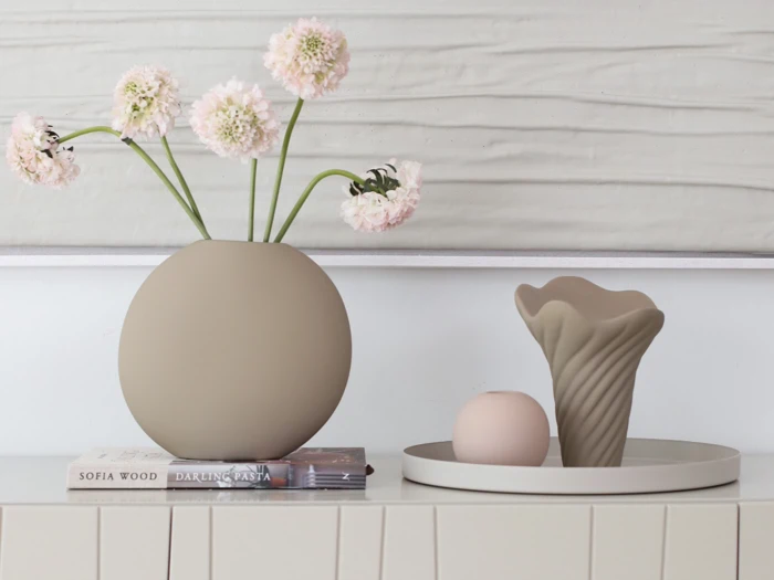 Rund vase med lyserøde blomster, en stak bøger og et bakke med en skulpturel vase og en kugle på et cremefarvet skab foran et vægmaleri med struktur.