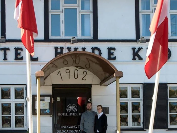 To personer står under en markise mærket 1902 ved indgangen til Hotel Hvide Kro med danske flag på hver side af døråbningen.