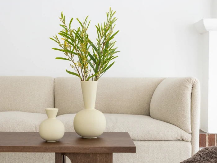 To cremefarvede vaser med grønne, bladrige grene står på et træbord foran en lysbeige sofa i en minimalistisk, moderne stue.