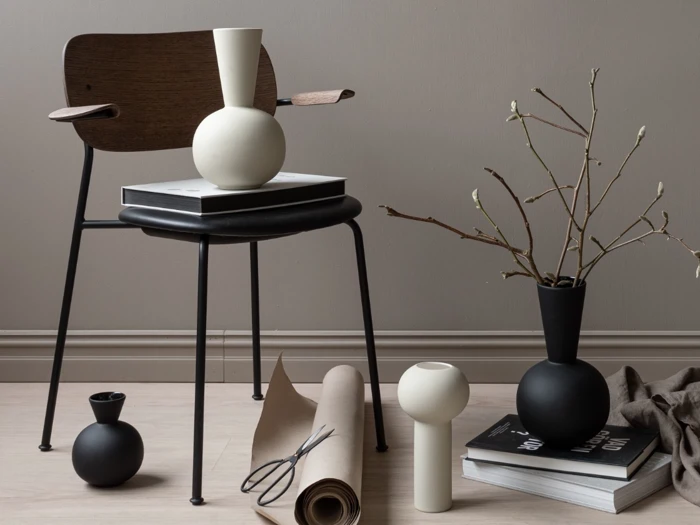 En moderne stol med bøger og vaser på og omkring den, et sammenrullet stykke papir og en gren i en vase, alt sammen placeret på et lyst trægulv foran en beige væg.