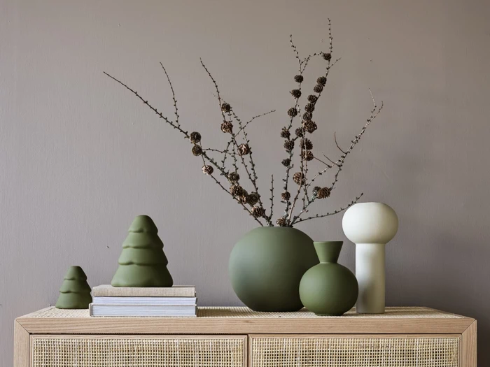 Minimalistisk indretning med grønne vaser, tørrede grene, en hvid abstrakt skulptur, to bøger og grønne træformede pyntegenstande på et træskab foran en taupefarvet væg.