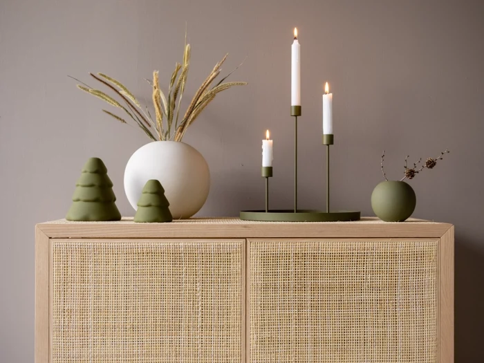 Minimalistisk indretning med en grøn lysestage, tændte stearinlys, en vase med tørret græs, to grønne juletræspynt og en lille rund vase på et kurveskab foran en taupefarvet væg.