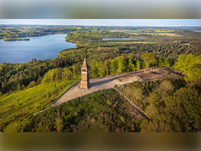Luftfoto af et højt murstenstårn på en bakke omgivet af grønne træer, med en sø og et skovlandskab i baggrunden.