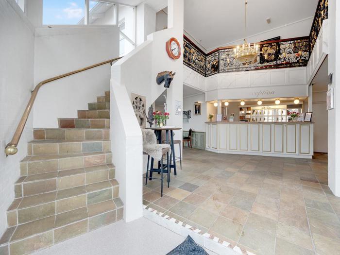 Lys hotellobby med klinkegulv, en trappe med messinggelænder, en receptionsskranke, højt til loftet og en stor lysekrone; dekorativ vægkunst over skranken.