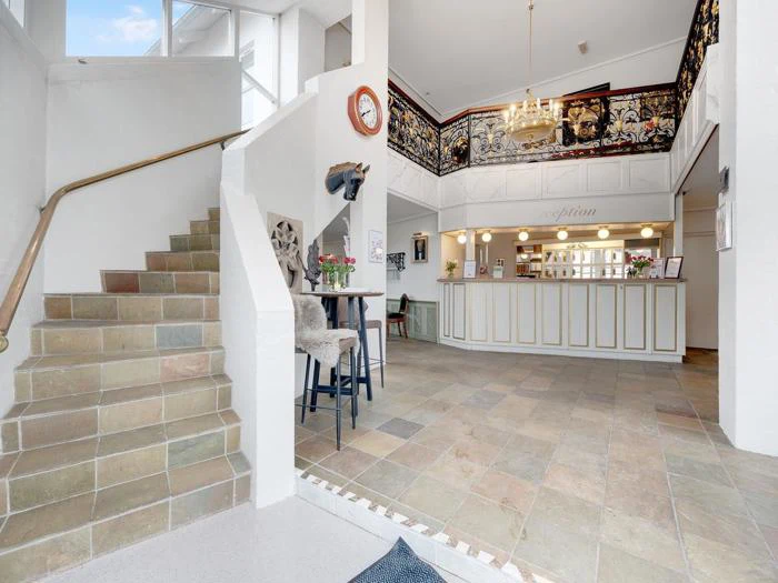 Rummelig hotellobby med klinkegulv, reception, lysekrone og trappe. Dekorativt gelænder og vægur tilføjer charme til det lyse, indbydende område.