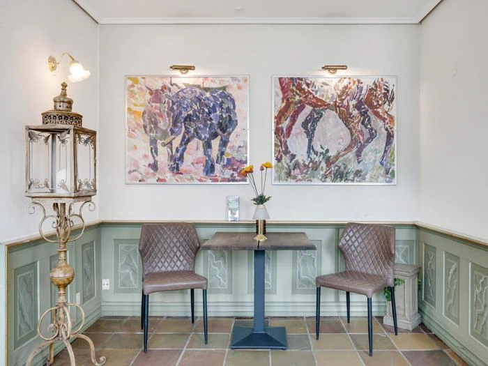 Et lille bord med to vatterede stole står under to farverige dyremalerier ved siden af en vintagelampe i et rum med grønne paneler og klinkegulv.